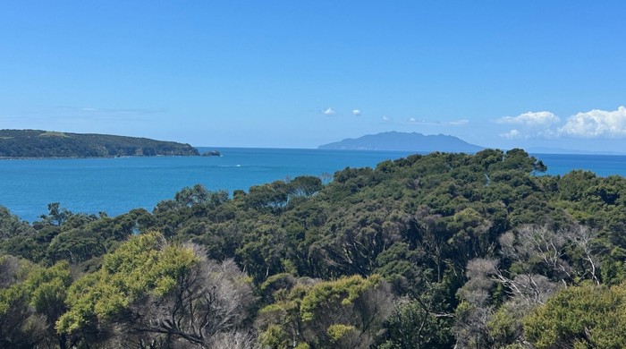 View from Kawau Island on a sunny day with a blue sky and a few clouds, with Hauturu / Little Barrier Island in the distance.