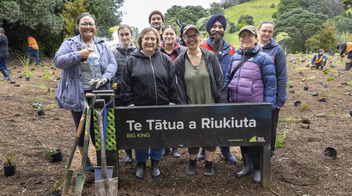 Nine people stand behind the main sign for the maunga. Four spades rest agains the sign and in the background people are planting small trees into the cleared ground,