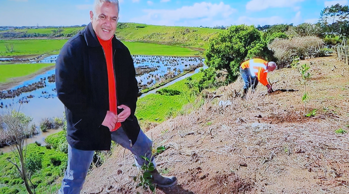 A man stands in the foreground smiling at the camera. Beneath him is a native tree seedling in the ground.