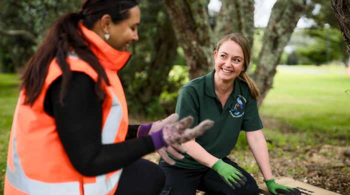 Two people outside under trees beside a pest animal trap in a wooden box on the ground.
