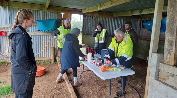 People in high-vis and rain jackets gather under a farm shelter around a table with snacks and drinks.